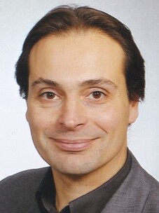 Porträt von Jochen Geck mit leichtem Lächeln und dunklem Hemd vor hellem Hintergrund.